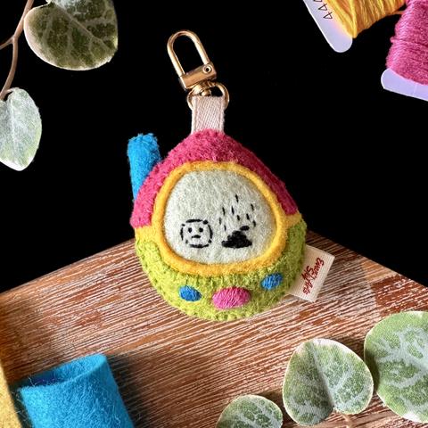 A product picture of a felt tamagotchi-like toys. It is egg-shaped, pink and green with embroidered buttons and a blue antenna. The creature embroidered on the screen is a sad blob next to a stinky poop. A ribbon linked to a golden lobster claw is attached to the top of it.