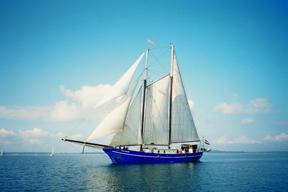 A blue sailing ship with multiple sails is anchored in calm waters under a clear blue sky, with a few clouds in the background.