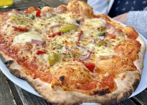 A close-up image of a freshly baked pizza topped with melted cheese, sliced red peppers, green peppers, and onions on a charred crust, served on a white plate. A drink is visible in the background.