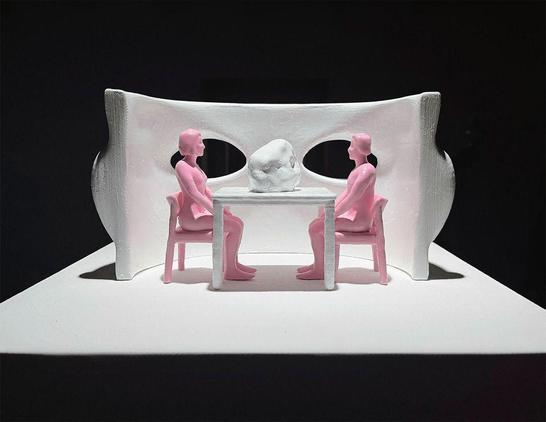 Two pink figures sit across from each other at a table with a rock in the center. A white sculptural backdrop frames the scene.
