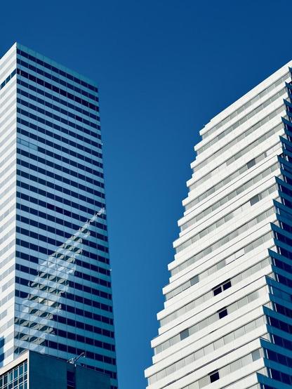 Two modern high-rise buildings against a clear blue sky, with one building reflecting the stepped facade of the other.