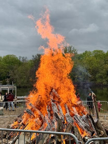 A large bonfire flames brightly with orange flames reaching upward, surrounded by wooden logs and a fenced area. In the background, people can be seen near a lake and trees under a cloudy sky.