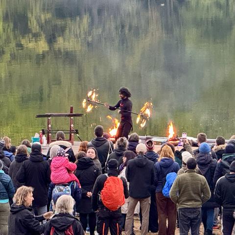 A fire performer showcases their skills on a platform, surrounded by an audience. The performance takes place near a body of water, with flames and reflections visible. The crowd, dressed in winter clothing, watches intently, some capturing the moment with phones.