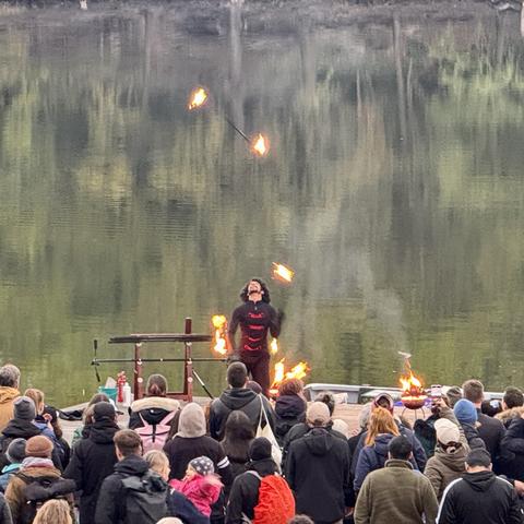 A fire performer juggles flaming objects in front of an audience by a lake. Spectators watch intently, with some taking photos. The performer wears a black outfit and displays a dramatic expression, surrounded by additional flames on the ground.