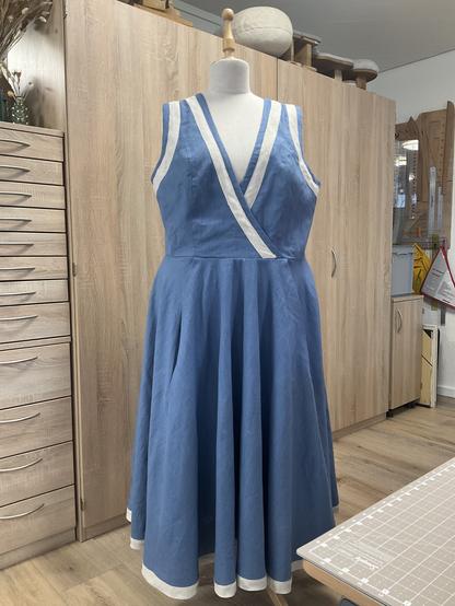 Stahlblaues Leinenkleid mit rohleinen Streifen an Hals- und Armausschnitten und a, Saum auf einer Schneiderpuppe vor einer Schrankwand.