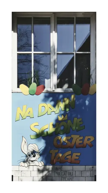Foto im Hochformat mit weißem breiten Rahmen, abgebildet in der oberen Bildhälfte eine zweiflügeliges Sprossenfenster, jeder Flügek ist vierfach geteilt. In der unteren Hälfte ist die Fassade hellblau gestrichen, am Fuss des Fensters sind übergroße Eier aus farbiger Pappe in gelb, grün und rot befestigt. Darunter steht in bunten Großbuchstaben auf vier Zeilen verteilt der Schriftzug 'NA DANN, SCHÖNE OSTERTAGE' links neben der Schrift auf Höhe der dritten und vierten Zeileein gezeichneter BugsBunny Hase der über eine ebenfalls aufgemalte Mauer schaut. Auf den weißen Mauerziegeln sthet weiter 'Euer Villa Rü Team'