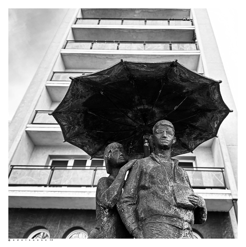 Schwarzweißfotografie: 3 Bronzefiguren (zwei nur sichtbar) unter einem Regenschirm, links eine junge Frau, rechts ein junger Mann mit angedeuteter Sonnenbrille und einem Buch, unterhalb der Brust haltend, im Hintergrund ein eckiges Hochhaus