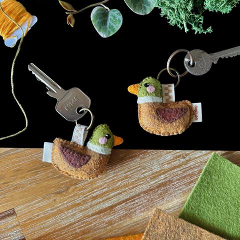 Two little handmade mallard duck keychains attached to keys and photographed on a black and wooden background, surrounded with decorative plants and matching felt and threads. 
The keychains are made of felt; they’re attached to a key ring with an ivory ribbon checkered with brown. 
The ducks are facing sideways, they have green heads, separated from their brown bodies with an ivory stripe. They have small embroidered black eyes and a little pink blush.