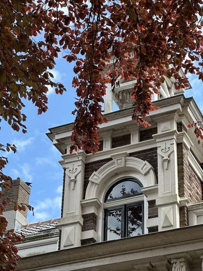 An architectural detail features a historic building with intricate white trim and a large arched window below a decorative tower. The scene is framed by branches with bright red and green leaves against a partly cloudy blue sky.