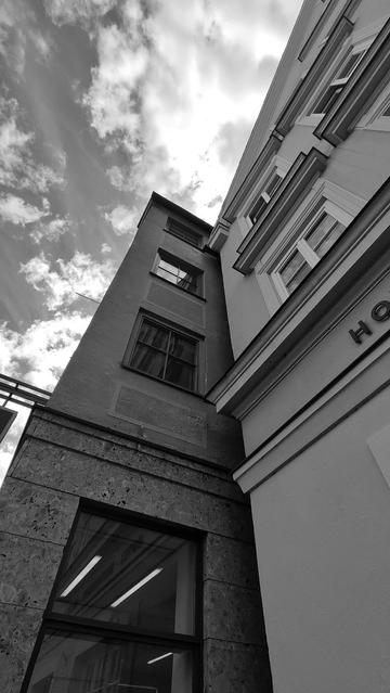Schwarz-weiß-Foto: Zwei zur Straße in der Tiefe versetzte Altstadtgebäude mit Sprossenfenstern in den oberen Etagen und einem Schaufenster im Erdgeschoss. Beim rechten Haus sind noch die Buchstaben "HO" zu sehen... vielleicht ein Hotel. Es ist ein sonniger Tag mit Wolken am Himmel.

Black and white photo: Two old town buildings offset from the street with lattice windows on the upper floors and a shop window on the ground floor. The letters "HO" can still be seen on the right-hand house... perhaps a hotel. It's a sunny day with clouds in the sky.