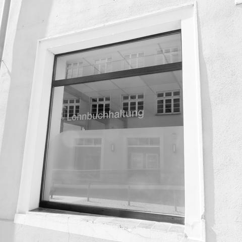 Schwarzweiß Foto.

Ein Fenster mit Oberlicht. 
Die Hälfte des unteren Bereiches ist mit Klebefolie  von innen blickdichter. 

Im Glasbereich steht 'Lohnbuchhaltung'.

Im Fenster spiegeln sich Fenster des gegenüberliegenden Hauses.