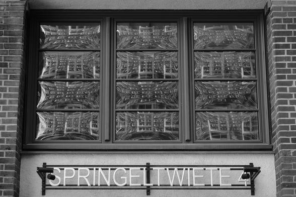 Blick auf ein dreigeteiltes Fenster mit vierfach geteilten gewölbten Scheiben, in denen sich die gegenüberliegende Backstein-Fassade mit vielen weiß eingefassten Fenstern spiegelt. Unten der Schriftzug Springeltwiete. Schwarz-weiß Photo.