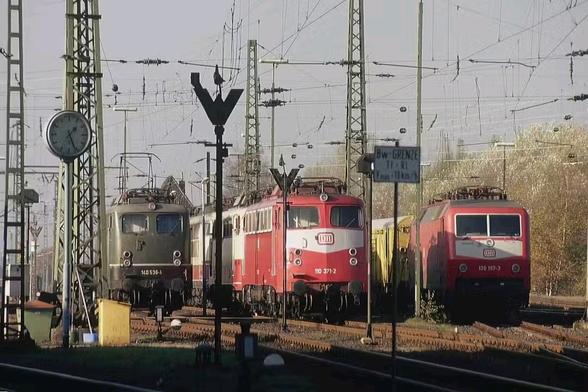 Ein lebendiges Bahnbetriebswerk der Deutschen Bundesbahn im Jahr 1990 zeigt drei nebeneinander stehende Elektrolokomotiven verschiedener Baureihen. Links steht eine grün lackierte Lok der Baureihe 140 mit der Betriebsnummer 140 536-4 im klassischen Bundesbahn-Stil der 1960er-Jahre. In der Mitte befindet sich eine rot-beige Lok der Baureihe 110 (Nummer 110 371-2) mit eleganter Stromlinienform – ein typischer Vertreter der Rheingold-Ära. Rechts daneben steht eine modern wirkende Lok der Baureihe 120 (Nummer 120 157-3) in verkehrsrotem Anstrich mit weißem Zierstreifen, der die technologische Zeitenwende in der Bahnindustrie einläutet.

Im Vordergrund befindet sich eine analoge Bahnhofuhr mit schwarzem Ziffernblatt auf weißem Grund. Umgeben sind die Loks von Oberleitungsmasten, Signalen, Kabeln und einem Schild mit der Aufschrift „Bw-GRENZE Tf-RL“, das die Grenze eines Bahnbetriebswerks markiert. Die Szene ist in weiches Sonnenlicht getaucht, das einen goldenen Herbstnachmittag andeutet. Das Bild dokumentiert eindrucksvoll den technischen Wandel zwischen Altbau- und Neubauloks im Übergang zur Deutschen Bahn AG.
