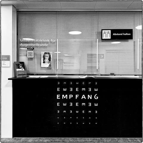 Quadratische Schwarzweissfotografie des Empfangsschalters einer Augenklinik, bei dem die Aufschrift "EMPFANG" umrahmt ist von einer "E-Haken"-Sehtafel.