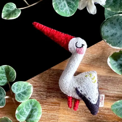 A little white stork felt plushie lying on a wooden and black background and surrounded with decorative plants.  The stork has a big red beak and small stick legs. Its design is angular, with a straight white neck and a round face, on which is embroidered an elegant and peacefully closed eye as well as a soft pink blush. Its body is triangular, with one of the corner covered with black felt to indicate the tip of the wings.  On its back, little leaves are embroidered.