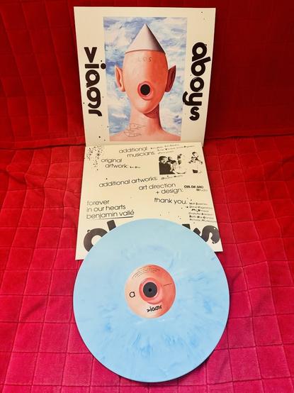 A vinyl record album cover for "Viagr aboys 🅴" by Viagra Boys is displayed. The cover art features a surreal painting of a bald figure with a cone hat, large ears, and a mouth replaced by a red circular speaker-like void. The word "LOS" is tattooed on the figure's forehead. The background is a bright sky with clouds. The vinyl itself is a light blue color with darker blue marbling, and the center label is pinkish-red. The album credits and artwork acknowledgments are also shown.