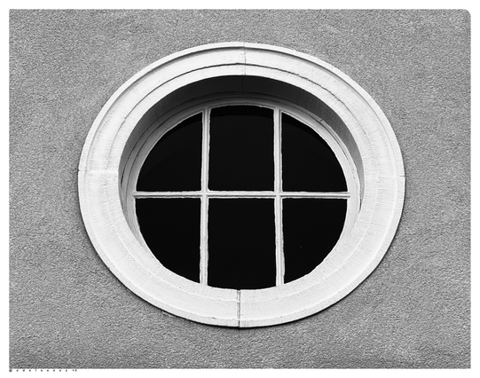 Schwarzweißfotografie: mittig ein kreisrundes Fenster, mit dunklen Fensterscheiben, weiße Streben und Rahmen, weiß eingefasst, ringsherum hellgrauer Rauputz