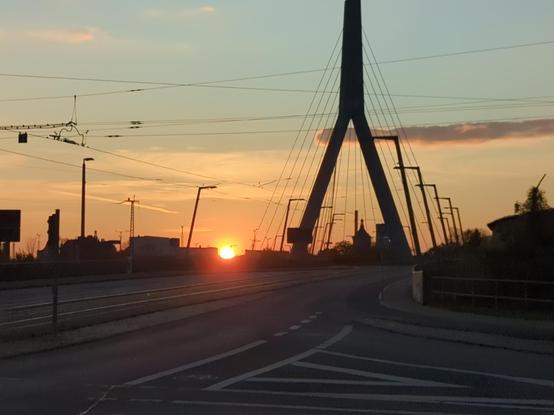 der Feuerball liegt rltlich golden genau zwischen zwei dachkanten am Horizont.
der Himmel fast wolkenlos. nur eine schmale Schleierwolke, die gerade de Pylon der Brücke küsst, deren Strassenbelag vollkommen leer ist.