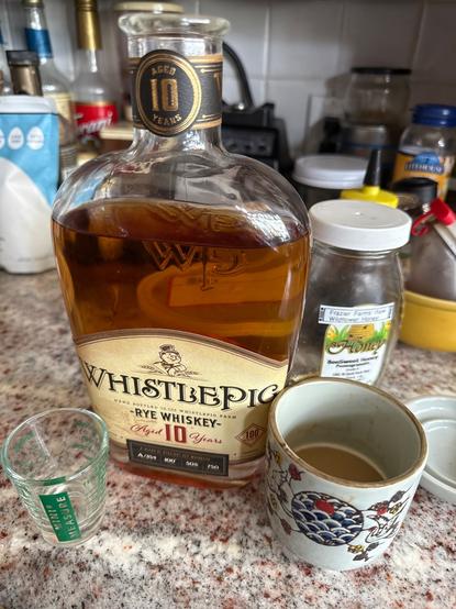 Adding 100 proof Whistlepig to the morning coffee. Mightn't be enough. There is a graduated cylinder shot glass, a Japanese ceramic cup, and a brown liquor in a flask bottle with a pig in a top hat logo siting on a speckled red and pink marble counter. The drink is half finished and likely needed per post.