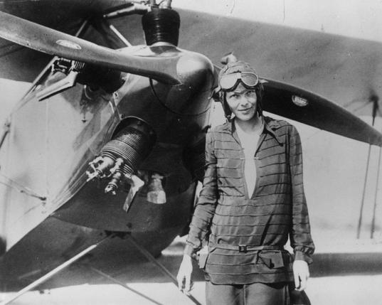 Amelia Earhart looking cool as fuck, with goggles and flight helmet, standing in front of a biplane.