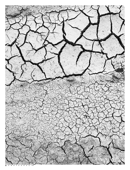 Schwarzweißfotografie: Blick vom oben auf ein Stück hellen Lehmboden, dieser ist ausgetrocknet und hat viele große und noch mehr kleinere Risse