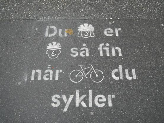 Auf der Straße ist mit weißer Farbe auf Norwegisch der Text aufgesprüht: "Du er så fin når du sykler" -"Du bist so schön wenn du radelst". Zwischen den Wörtern sind als Symbole Köpfe mit Fahrradhelmen und ein Fahrrad zu sehen.