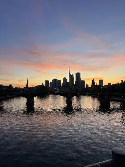 Die Skyline Frankfurt im Sonnenuntergang von der Flößerbrücke Frankfurt aus. Der Himmel ist orange gelb und spiegelt sich farblich ein wenig im Fluss.