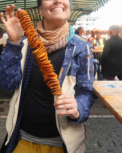 Alex hält lachend Essen in die Kamera: Eine aufgeschnittene, evrdrehte Kartoffel an einem langen Spieß.