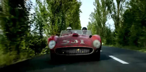 A still from the film Ferrari. A classic red sports car with the numbers 531 painted on its bonnet speeds along a country road. The trees to either side are blurred.