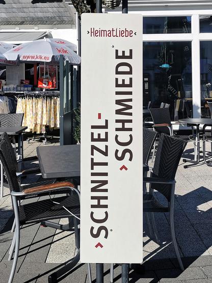Personenhoher Aufsteller vor einigen Läden. Senkrecht groß: "Schnitzel-Schmiede", darüber klein horizontal "Heimatliebe"