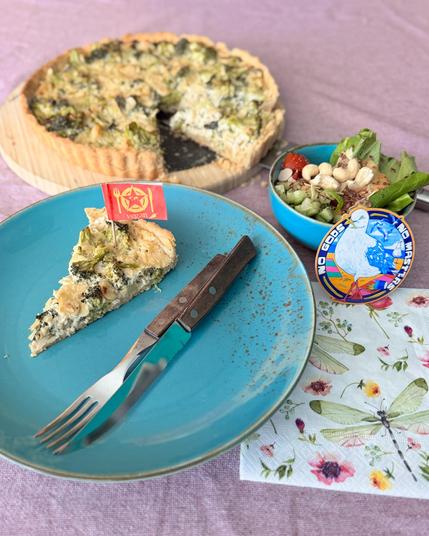 Das Bild zeigt eine appetitliche Mahlzeit auf einem lilafarbenen Tisch. Im Vordergrund befindet sich ein blauer Teller mit einem Stück veganer Quiche, das mit einer kleinen roten Fahne mit dem Wort "vegan" und einem Symbol versehen ist. Neben dem Teller liegt eine Gabel und ein Messer. Im Hintergrund ist eine Quiche auf einem Holztablett zu sehen, aus der ein Stück fehlte. Neben der Quiche steht eine blaue Schüssel mit einer Salatmischung, die aus grünen Blättern, Tomaten, Nüssen und anderen Zutaten besteht. Ein buntes Etikett mit der Aufschrift "NO GODS NO MASTERS" und einer weißen Möwe ist ebenfalls sichtbar. Eine Serviette mit Blumen und Libellen liegt unter dem Teller.