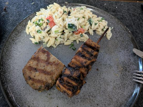 Ein grauer runder Teller, auf der unteren Hälfte links ein halbes Steak, rechts daneben der planted Grillspieß, auf der oberen Tellerhälfte Nudelsalat