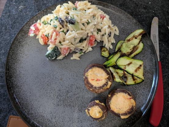 Der gleiche graue Teller, auch mit Nudelsalat auf der oberen Hälfte, auf de unteren rechts 3 gefüllte Champignons und ein paar Scheiben gegrillte Zucchini. Neben dem Teller rechts liegt ein Messer mit rotem Griff