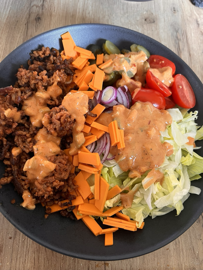 Das Foto zeigt einen Teller mit veganer Cheeseburger Bowl. Sie besteht aus veganem Hackfleischersatz, veganem Cheddar, Salat, Zwiebeln, Tomaten und Essiggurten.