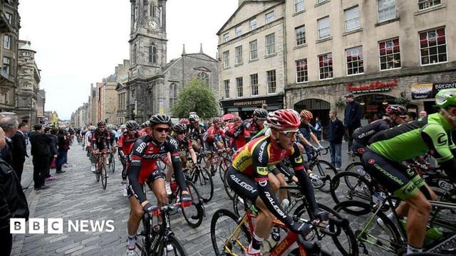 A group of cyclists, known as a peleton, travelling from left to right on the Royal Mile in Edinburgh. There are people watching by the side of the road. A Starbucks and the Tron Church can be seen in the background.