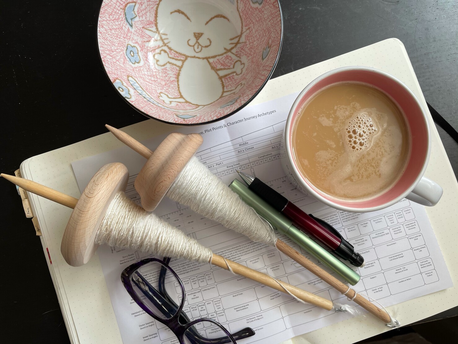 Foto von oben auf mein Bulletjournal, darauf die Lesebrille, zwei „vollen“ Handspindeln, Füller und Bleistift, eine gefüllte Kaffeetasse und eine Schale mit einer gezeichneten Comic-Katze auf dem Boden Foto von oben auf mein Bulletjournal, darauf die Lesebrille, zwei „vollen“ Handspindeln, Füller und Bleistift, eine gefüllte Kaffeetasse und eine Schale mit einer gezeichneten Comic-Katze auf dem Boden