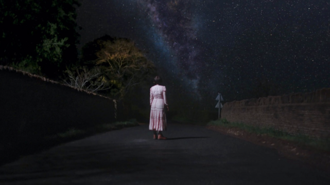 »Men«-Standbild. Jessie Buckley allein auf einer Dorfstraße in der Nacht, vor ihr die Milchstraße.
