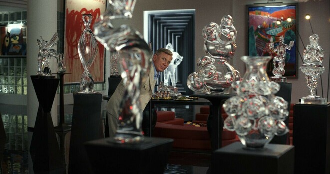 »Glass Onion. A Knive’s Out Mystery«-Standbild. Daniel Craig prüft als Benoit Blanc Projektil-Linie umgeben von kitschigen Glass-Skulpturen.