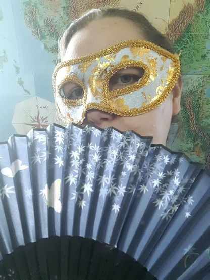 Ein  Selfie von mir, ich trage eine weiß-goldene venezianische Maske und halte einen blauen Fächer mit weißen Blumen und Schmetterlingen vor mein Gesicht. Im Hintergrund sieht man eine Aventurien-Karte.