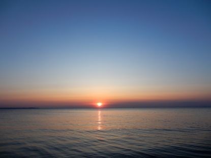 Sonnenuntergang über dem Meer: Blauer Himmel nimmt die oberen zwei Drittel ein, darunter das spiegelglatte Meer. In der Mitte die Sonne direkt am Horizont, leichte rötliche Färbung trennt Meer und Himmel.