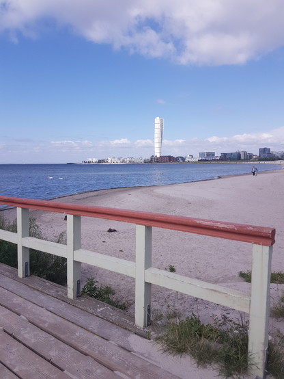 Im Hintergrund ist ein gedrehter Turm umrahmt von blauem Himmel. Im Vordergrund eine hölzerne Brücke. Dazwischen sind Strand und Meer. 