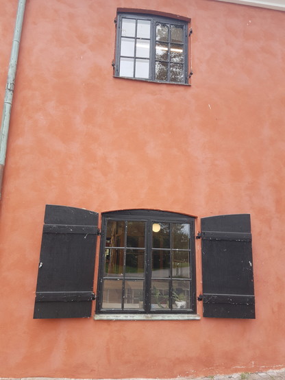 Orangene Hauswand mit schwarzen Fensterrahmen. Das untere Fenster hat schwarze, aufgeklappte Fensterläden. 