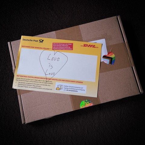Kleines Päckchen mit einem herzförmigen Pride-/Regenbogenaufkleber und eine DHL-Benachrichtigungskarte, auf die 