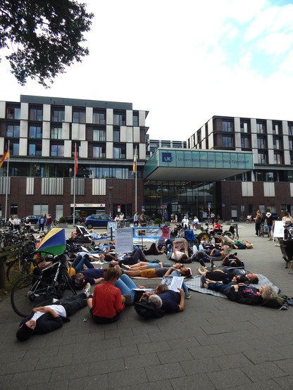 Vor dem UKE Hamburg-Eppendorf liegt eine Gruppe Menschen mit Protestschildern auf dem Boden.