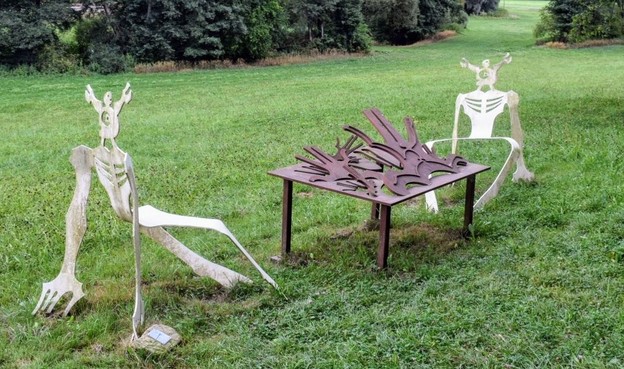 Skulptur "Frühstück im Freien" auf einem Wanderweg bei Waldenbuch. Zwei dünne, weiße skelettartige Figuren sitzen auf der Wiese an einem metallenen Tisch, des Tischplatte in großen Splittern aufwölbt. 