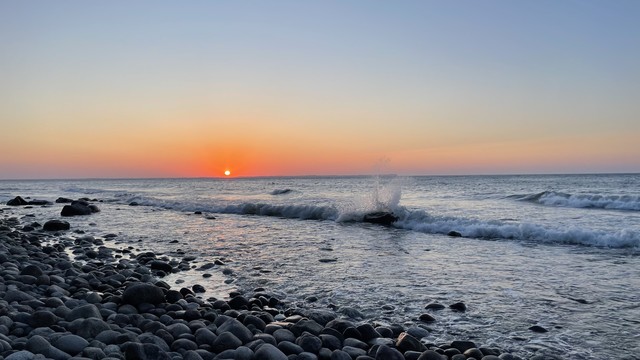 Foto des Sonnenuntergangs am Meer. Links im Bild ein Steinstrand mit großen, grauen Steinen. Im flachen Uferbereich liegen große dunkle Steine. Das Meer rollt Wellen in gleichmäßigen Abständen Richtung Ufer. Knapp über dem Horizont links der Bildmitte hängt die kleine, runde, orange leuchtende Sonne. Der wolkenlose Himmel um sie herum in kräftigen Gelb-, Orange- und Rottönen. In Ufernähe bricht sich eine Welle über einem großen, dunklen Stein und wirft Wasser nach oben. 