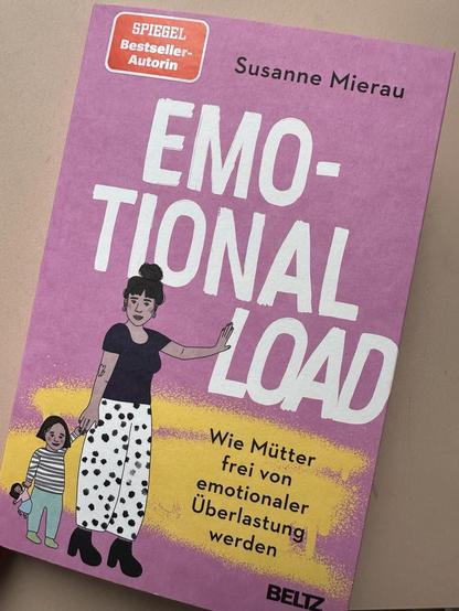 Foto von Buchcover: Emotional Load von Susanne Mierau. Rosa Cover mit weißem Schriftzug. Untertitel auf gelber Sprühfarbe: Wie Mütter frei von emotionaler Überlastung werden. Auf dem Cover befindet sich eine Illustration von Johanna Augustin (Konfettikrake): Mutter mit Kind an der Hand schiebt das Wort „Load“ weg
