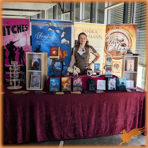 Foto meines Stands an einer Fensterfront mit gegen direkte Sonne runtergelassenen Jalousien. Am Stand stehen Bücher,cLesezeichen, Tassen und Schlüsselanhänger plus etwas Deko. Ich (Frau Ende 20, lange dunkelblonde Haare) trage ein Fantasy-Ledermieder. 